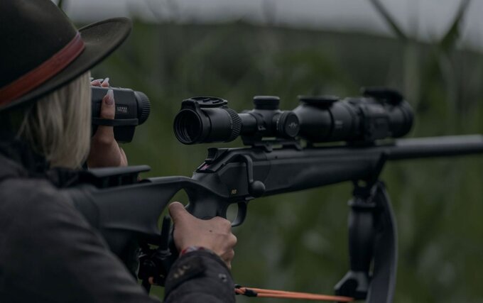Person in Outdoor-Kleidung beobachtet mit einem Handgerät das Umfeld, während ein Gewehr mit Zielfernrohr auf einer Auflage ausgerichtet ist; Aufnahme im Freien, thematisch Jagd und Nachtsichttechnik.