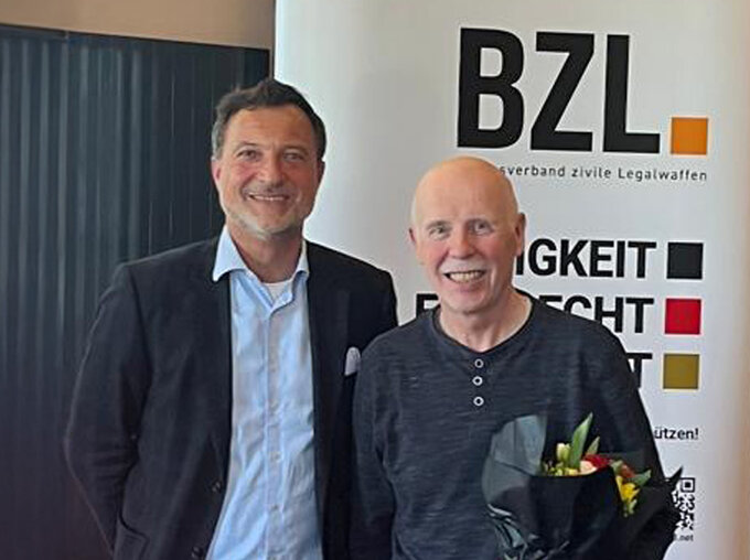 Matthias Klotz, Vorsitzender des Bundesverbands zivile Legalwaffen e. V. (BZL), verabschiedet Walter Finke vom BHDS (rechts), der aus der aktiven Verbandsarbeit ausscheidet, und überreicht ihm ein kleines Präsent als Dank für die Zusammenarbeit; im Hintergrund ein BZL-Roll-up.