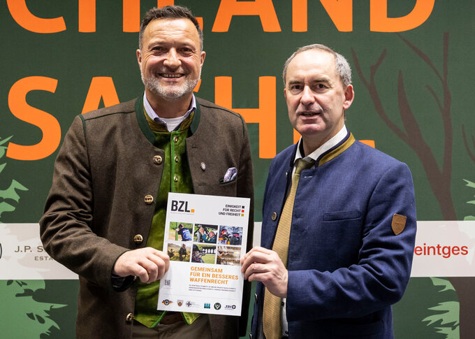 Zwei Männer stehen nebeneinander bei einer Veranstaltung und halten gemeinsam eine Broschüre in die Kamera. Beide tragen traditionelle Jacken, im Hintergrund ist eine beschriftete Veranstaltungswand zu sehen.