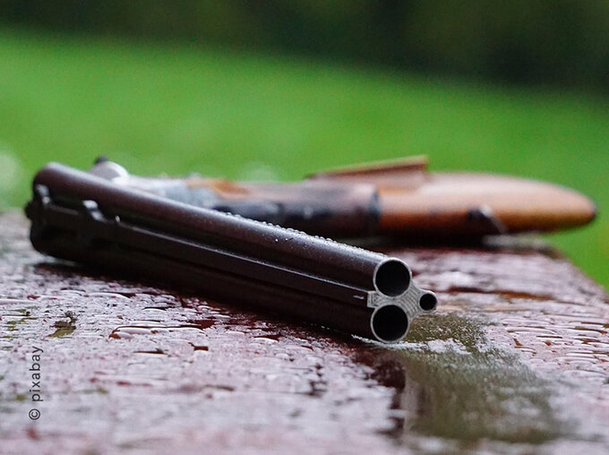 Nahaufnahme der Läufe einer Doppelflinte, die auf einer feuchten Holzoberfläche im Freien liegt; unscharfer grüner Hintergrund, symbolisches Bild zum Thema Jagd und Waffenrecht.