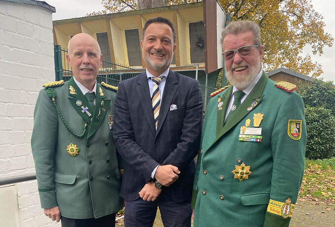 Drei Männer stehen im Freien vor einem Gebäude; zwei tragen grüne Schützenuniformen mit Orden und Abzeichen, der Mann in der Mitte einen dunklen Anzug mit Krawatte. Gruppenfoto bei einem Vor-Ort-Termin im schießsportlichen Umfeld.