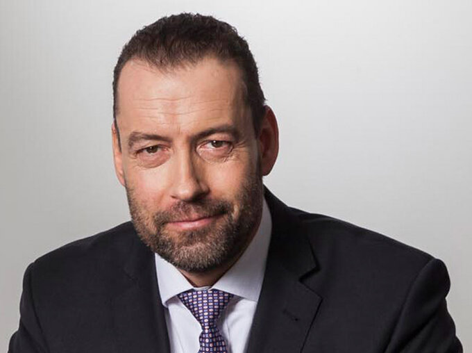 Porträt eines Mannes mit Bart im dunklen Anzug und Krawatte vor hellem, neutralem Hintergrund; professionelles Pressefoto eines Politikers.