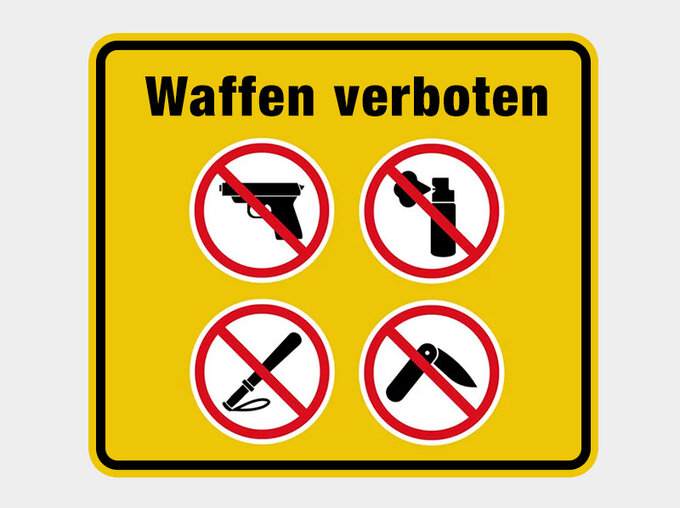 Gelbes Hinweisschild mit der Aufschrift „Waffen verboten“ und vier durchgestrichenen Piktogrammen für Pistole, Reizgas, Messer und Schlagstock.