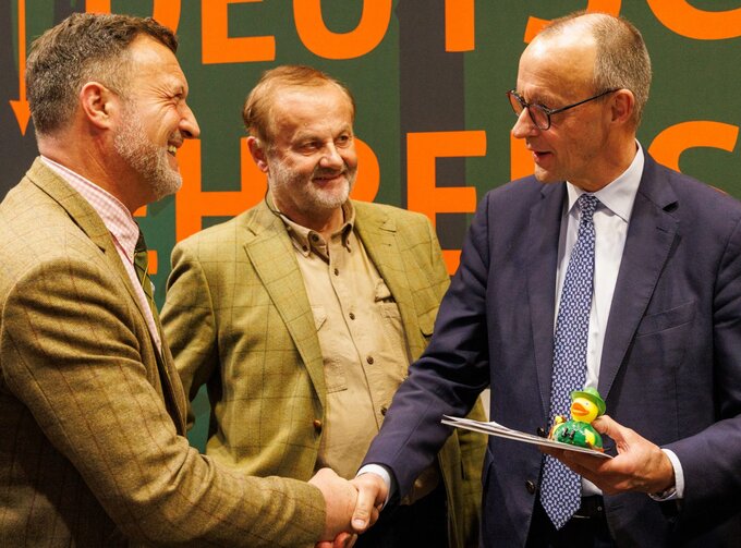 Drei Männer im Gespräch, zwei von ihnen geben sich die Hand. Einer hält Unterlagen und eine kleine Spielfigur, im Hintergrund ist eine Messe- oder Veranstaltungswand zu sehen. Unter anderen Bundeskanzler Friedrich Merz (rechts)