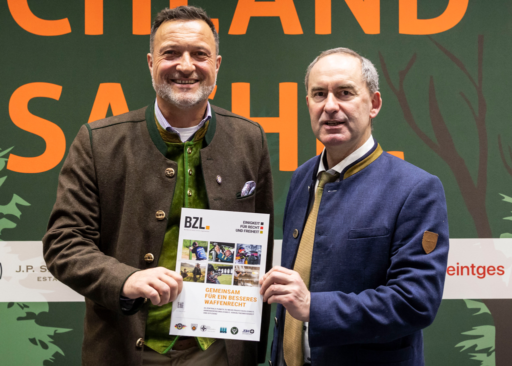 Zwei Männer stehen nebeneinander bei einer Veranstaltung und halten gemeinsam eine Broschüre in die Kamera. Beide tragen traditionelle Jacken, im Hintergrund ist eine beschriftete Veranstaltungswand zu sehen.