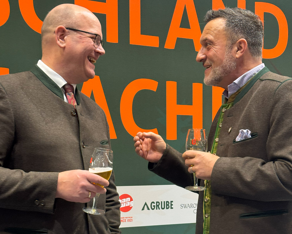 Zwei Männer stehen im Gespräch bei einer Veranstaltung und halten jeweils ein Glas Getränk in der Hand. Sie lachen und unterhalten sich vor einer beschrifteten Messe- oder Veranstaltungswand.