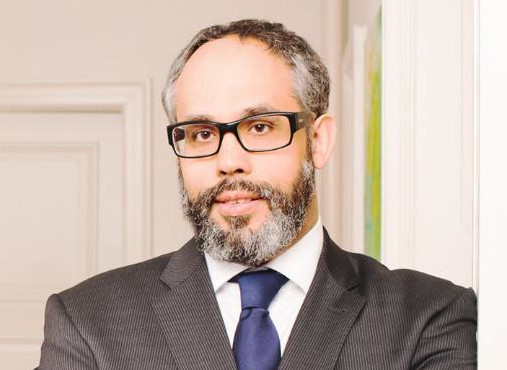 Porträt eines Mannes mit Brille und Bart im dunklen Anzug und Krawatte vor hellem Innenraum; professionelles Pressefoto eines Politikers.